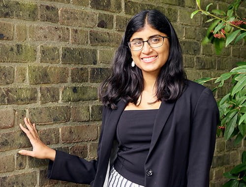 ACA student Ashwini Poopalasingham young asian woman standing in front of a brick wall and tree black clothing long hair