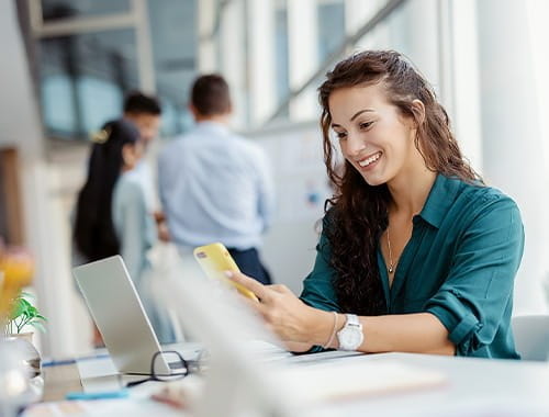 young woman employee working in an office smiling looking at mobile phone laptop desk