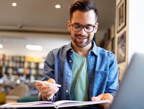 young man glasses student studying pen paper folder book denim shirt laptop office library