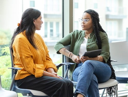 two women colleagues businesswomen having a difficult conversation in an office meeting room sat on white chairs in front of windows