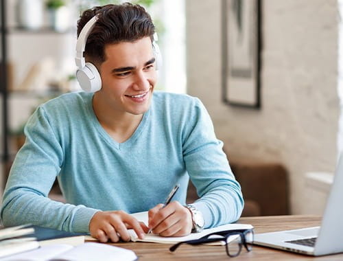 young man student studying looking at laptop wearing headphones writing taking notes