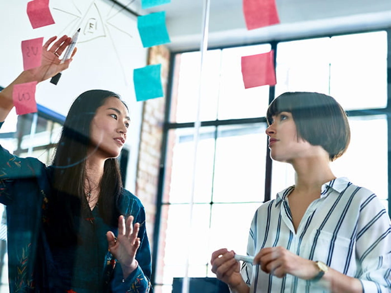 two women colleagues brainstorming meeting office large windows glass post it notes