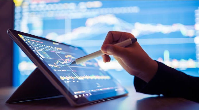 A hand working on a tablet showing graphs, with a larger screen also showing graphs in the background