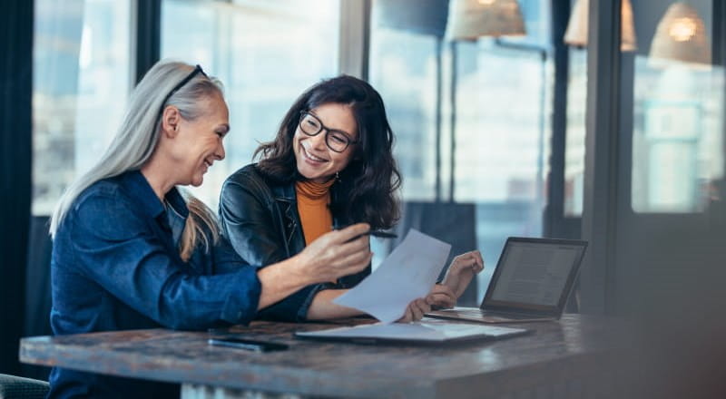 Mature woman advising younger woman at desk in glass office
