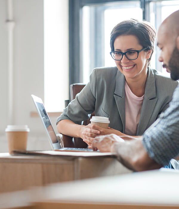 Business person working with a colleague at a laptop smiling, holding a cup of coffee