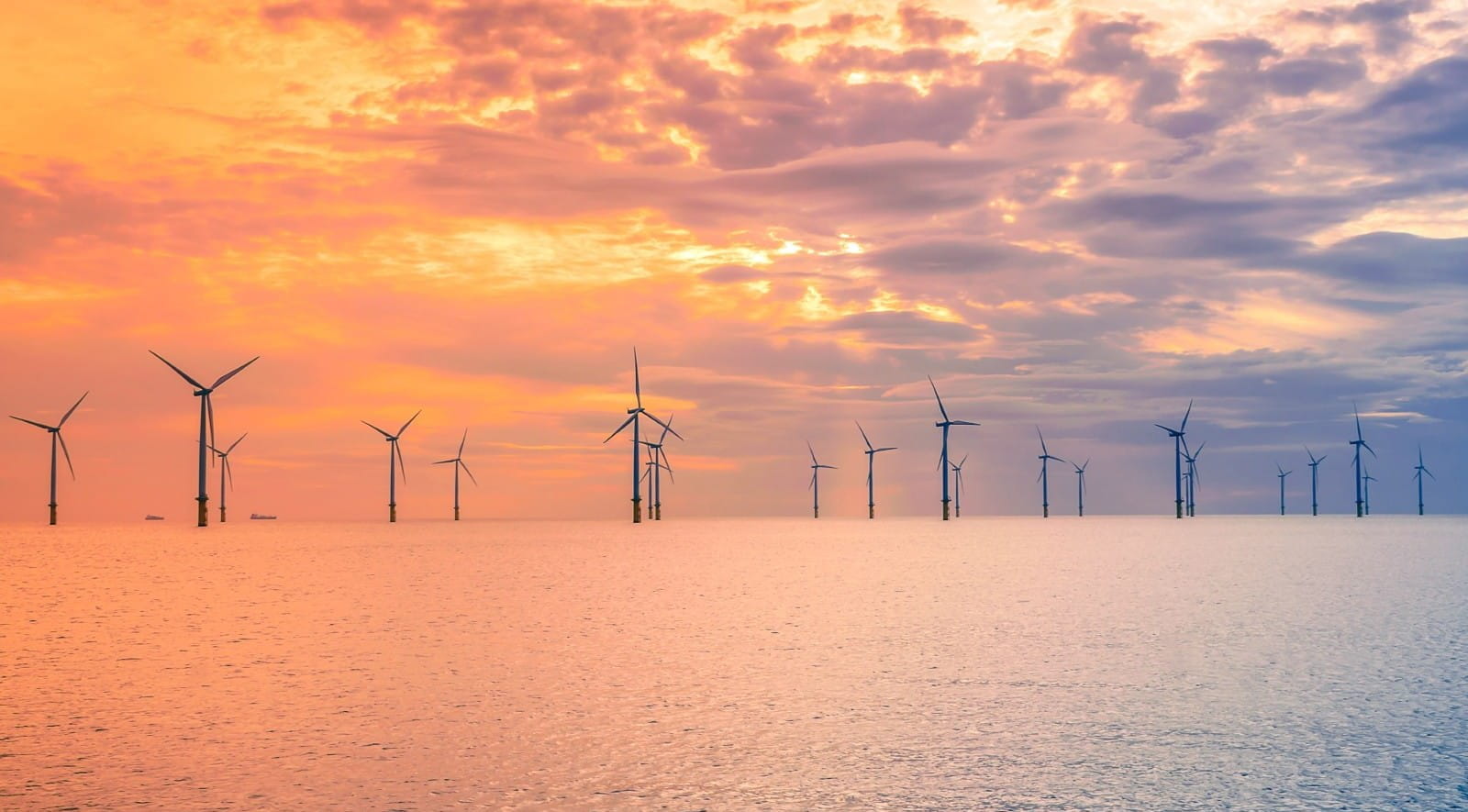 Four wind turbines in the countryside with hills in the distance, and the sun beginning to set behind them.
