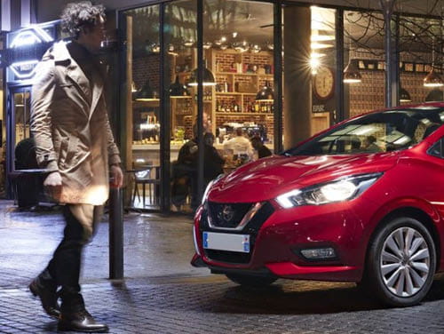 A person walking by a red car at night with a bar in the background